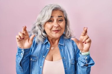 Middle age woman with grey hair standing over pink background gesturing finger crossed smiling with hope and eyes closed. luck and superstitious concept. 