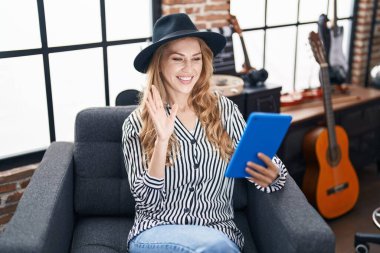 Beautiful blonde woman at music studio using tablet looking positive and happy standing and smiling with a confident smile showing teeth 