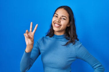 Young brazilian woman standing over blue isolated background smiling looking to the camera showing fingers doing victory sign. number two. 