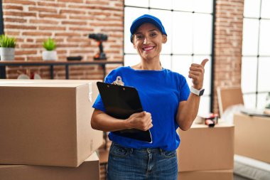 Middle age brunette woman wearing delivery uniform at house moving smiling happy and positive, thumb up doing excellent and approval sign 