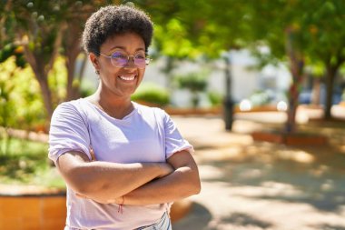 Afro-Amerikalı kadın gülümsüyor. Parkta kollarını kavuşturmuş duruyor.