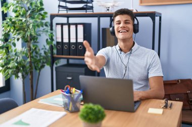 Young hispanic man working at the office wearing headphones smiling friendly offering handshake as greeting and welcoming. successful business. 