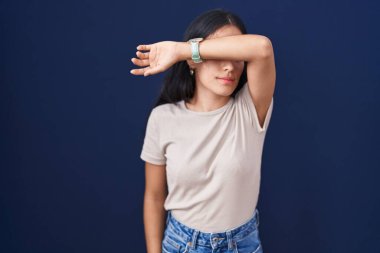 Mavi arka planda dikilen genç İspanyol kadın kollarıyla gözlerini kapatıyor, ciddi ve üzgün görünüyor. Görme, saklama ve reddetme konsepti 