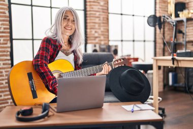 Müzik stüdyosunda dizüstü bilgisayar kullanarak klasik gitar çalan gri saçlı kadın müzisyen.
