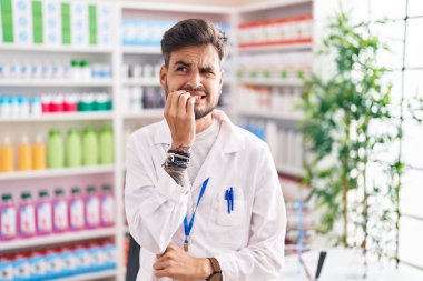 Young hispanic man with tattoos working at pharmacy drugstore looking stressed and nervous with hands on mouth biting nails. anxiety problem. 