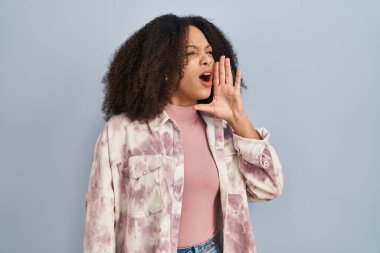 Young african american woman standing over blue background shouting and screaming loud to side with hand on mouth. communication concept. 