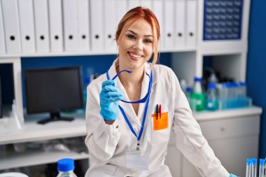 Beyaz kadın bilim adamı gülümsüyor. Laboratuvarda kendine güvenen bir gözlük tutuyor.