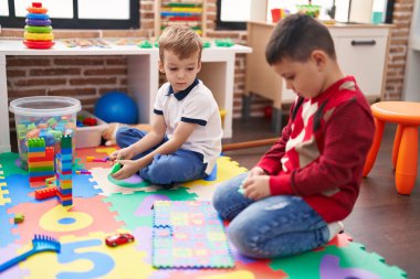 Anaokulunda yerde oturmuş, inşaat blokları ve matematik bulmacalarıyla oynayan iki çocuk.