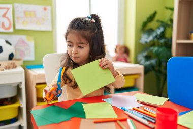 Adorable hispanic girl student sitting on table cutting paper at kindergarten