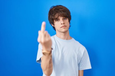 Hispanic young man standing over blue background showing middle finger, impolite and rude fuck off expression 