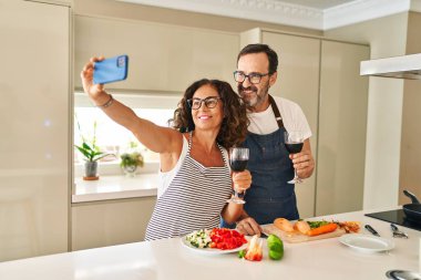 Middle age hispanic couple smiling confident cooking and make selfie by the smartphone at kitchen
