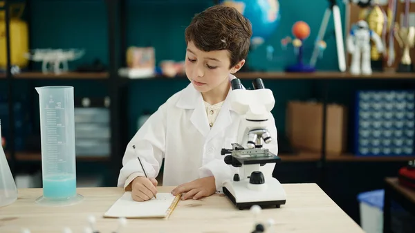 Adorable Hispanic Boy Student Using Microscope Writing Notebook Laboratory Classroom — 图库照片