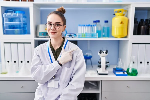 Young hispanic girl working at scientist laboratory smiling cheerful pointing with hand and finger up to the side