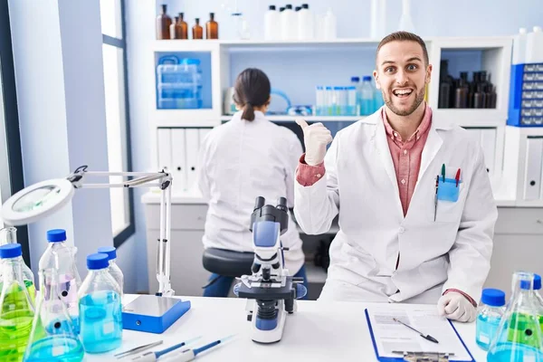 Young man working at scientist laboratory pointing thumb up to the side smiling happy with open mouth