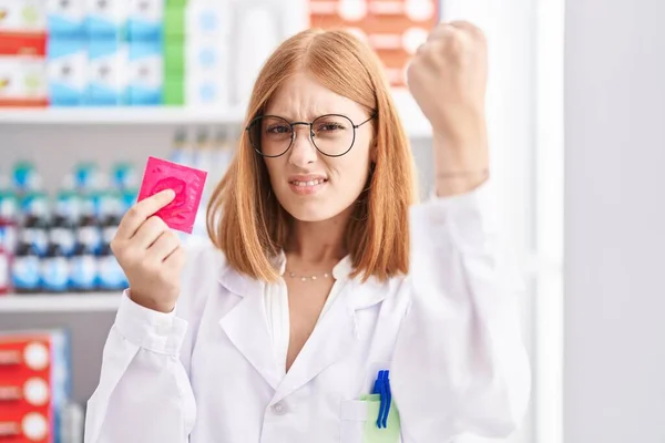 Young Redhead Woman Working Pharmacy Drugstore Holding Condom Annoyed Frustrated — Zdjęcie stockowe