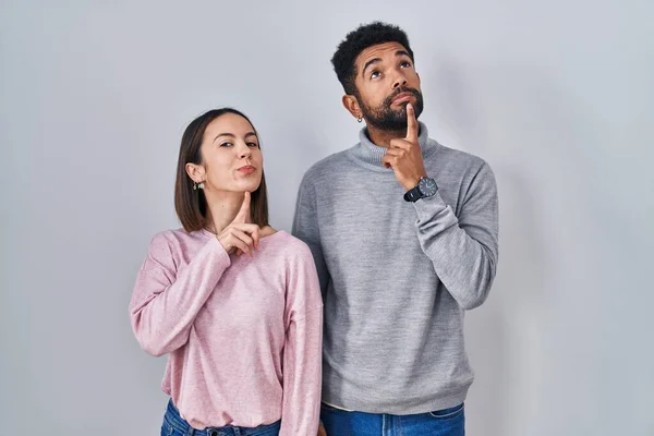 Young hispanic couple standing together thinking concentrated about doubt with finger on chin and looking up wondering