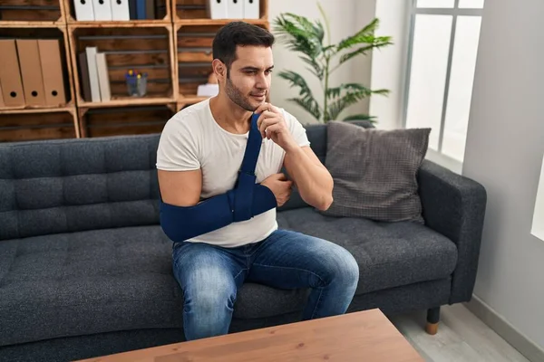 Young hispanic man with beard wearing arm on sling sitting at therapy consult serious face thinking about question with hand on chin, thoughtful about confusing idea 