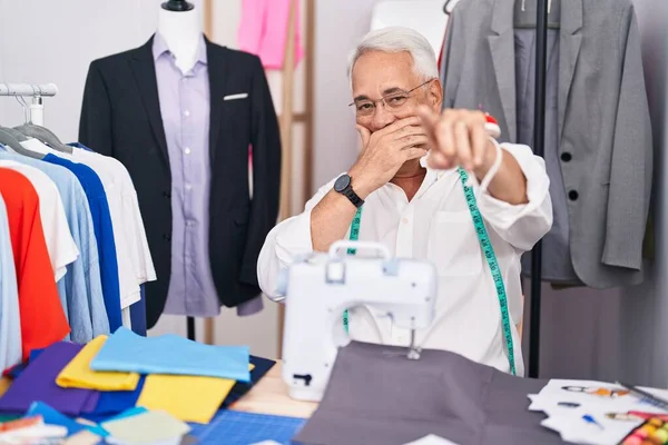 Middle age man with grey hair dressmaker using sewing machine laughing at you, pointing finger to the camera with hand over mouth, shame expression