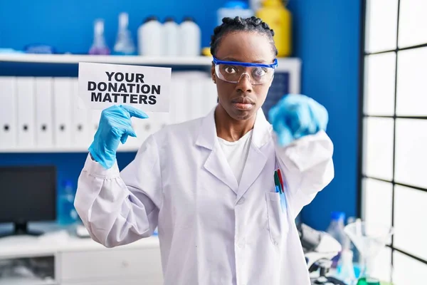 Beautiful black woman working at scientist laboratory holding your donation matters banner pointing with finger to the camera and to you, confident gesture looking serious