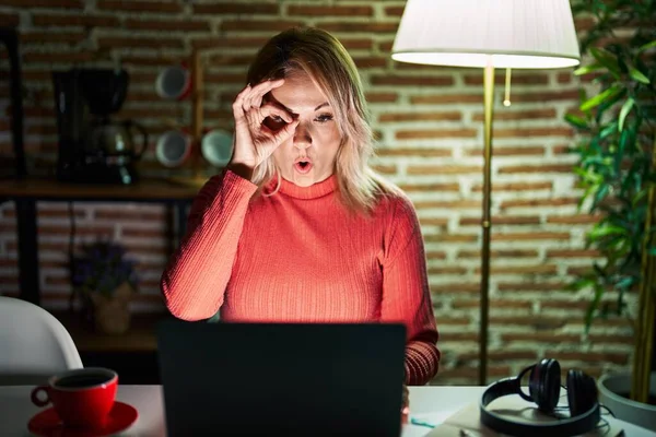 Blonde woman using laptop at night at home doing ok gesture shocked with surprised face, eye looking through fingers. unbelieving expression.