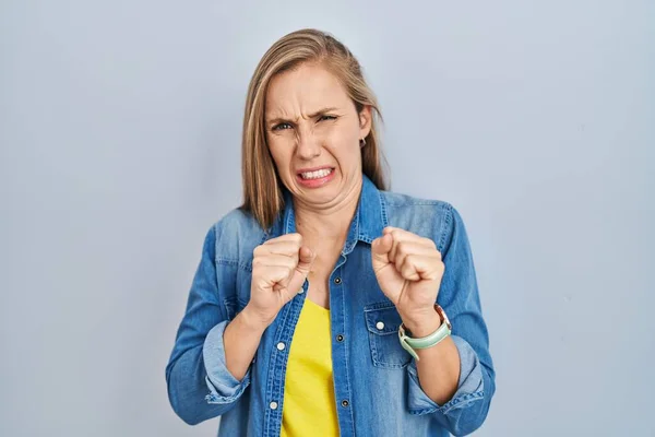 Young blonde woman standing over blue background disgusted expression, displeased and fearful doing disgust face because aversion reaction.