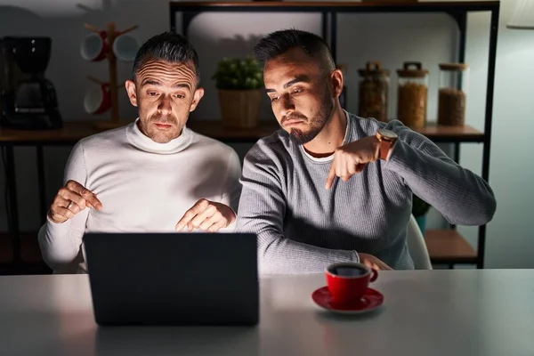 Homosexual couple using computer laptop pointing down looking sad and upset, indicating direction with fingers, unhappy and depressed.