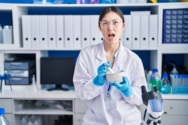 Chinese young woman working at scientist laboratory mixing angry and mad screaming frustrated and furious, shouting with anger looking up.