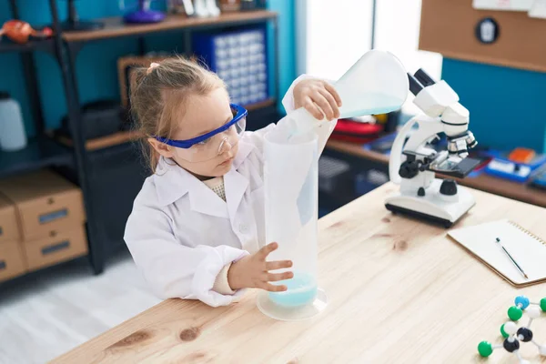 Adorable blonde girl student pouring liquid on test tube at laboratory classroom
