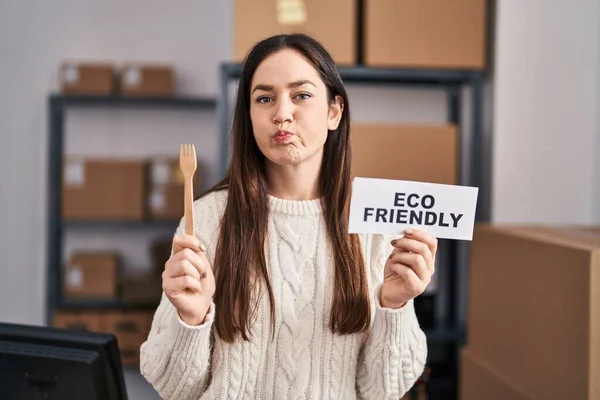 Young brunette woman working at eco friendly ecommerce puffing cheeks with funny face. mouth inflated with air, catching air.