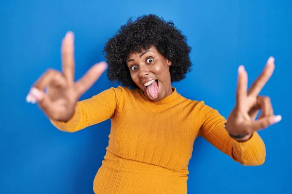 Black woman with curly hair standing over blue background smiling with tongue out showing fingers of both hands doing victory sign. number two.
