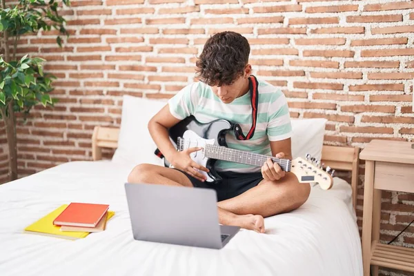 Young hispanic teenager having online electrical guitar class sitting on bed at bedroom