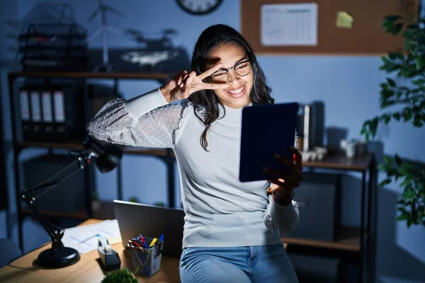 Young brazilian woman using touchpad at night working at the office doing peace symbol with fingers over face, smiling cheerful showing victory