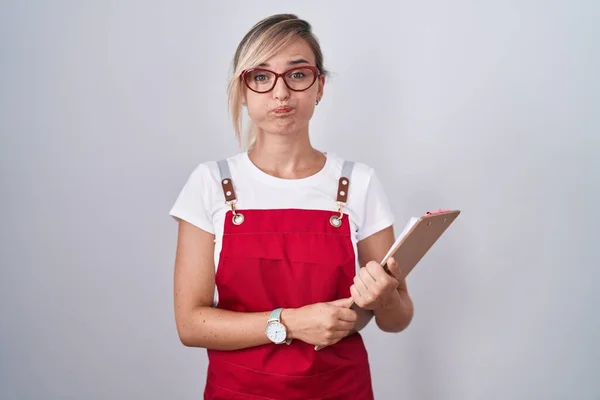 Young Blonde Woman Wearing Waiter Uniform Holding Clipboard Puffing Cheeks — Stockfoto