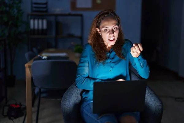 Brunette woman working at the office at night angry and mad raising fist frustrated and furious while shouting with anger. rage and aggressive concept.