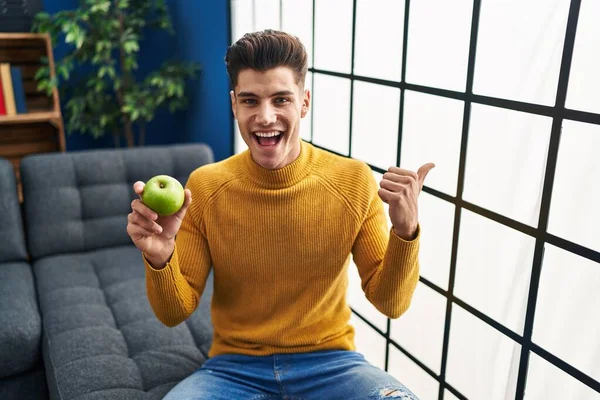 Young Hispanic Man Holding Green Apple Pointing Thumb Side Smiling — Zdjęcie stockowe