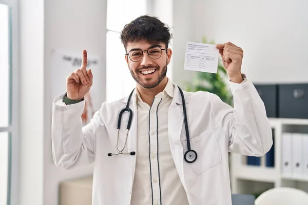 Hispanic doctor man with beard holding covid record card surprised with an idea or question pointing finger with happy face, number one