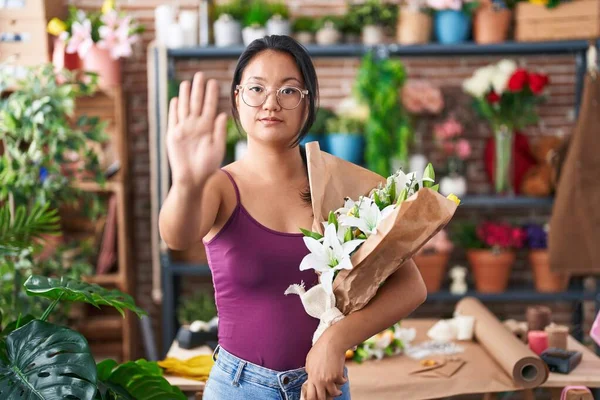 Asian young woman at florist shop holding bouquet of flowers with open hand doing stop sign with serious and confident expression, defense gesture