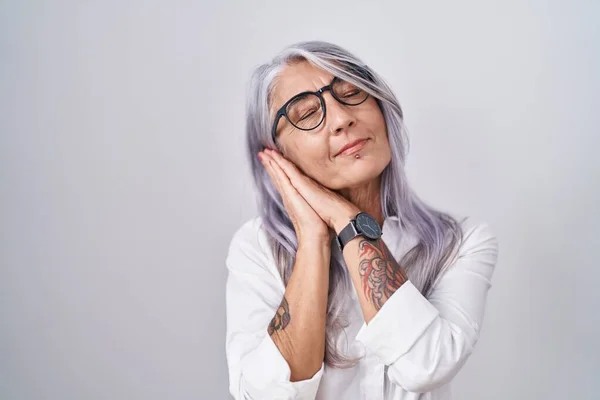Middle age woman with tattoos wearing glasses standing over white background sleeping tired dreaming and posing with hands together while smiling with closed eyes.