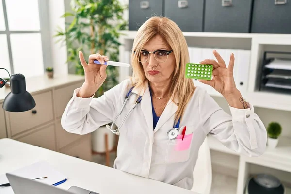 Middle age blonde doctor woman holding prescription pills and pregnancy test skeptic and nervous, frowning upset because of problem. negative person.