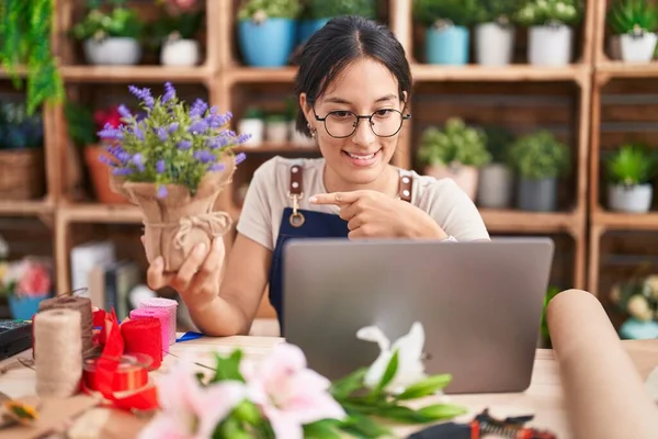 Young hispanic woman working at florist shop doing video call pointing to the back behind with hand and thumbs up, smiling confident