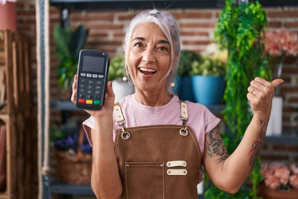 Middle age woman with tattoos working at florist shop holding dataphone pointing thumb up to the side smiling happy with open mouth