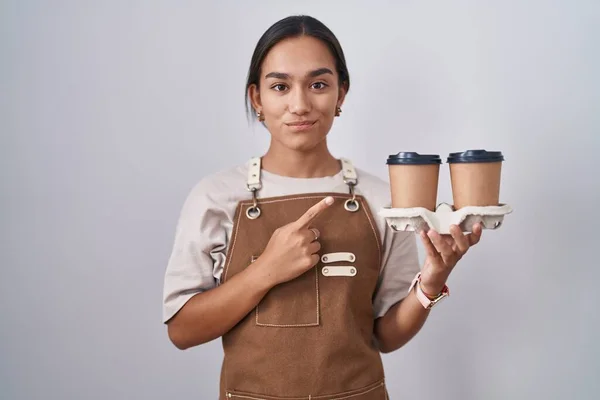 Young hispanic woman wearing professional waitress apron holding coffee pointing with hand finger to the side showing advertisement, serious and calm face
