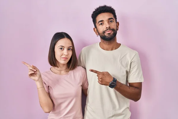他的年轻夫妇在粉色的背景上手指头指向广告的侧面 严肃而沉着的面孔 — 图库照片