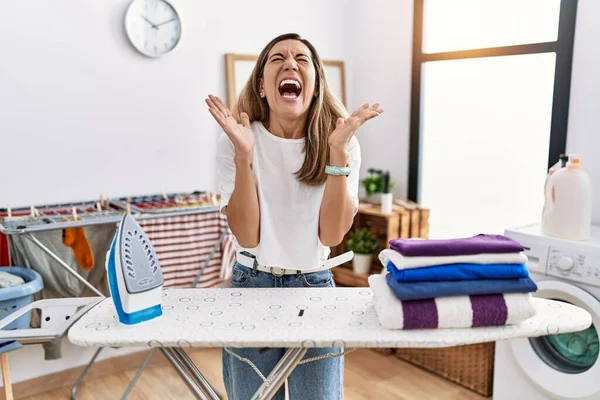 腕を上げ 興奮叫んで目を閉じて成功のために狂気と狂気を祝う洗濯室で若いヒスパニック系の女性のアイロン服 勝者のコンセプト — ストック写真