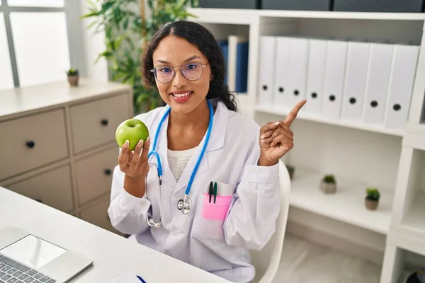 年轻的惊慌失措的医生女人拿着绿色的苹果开心地微笑着 手指头指向旁边 — 图库照片