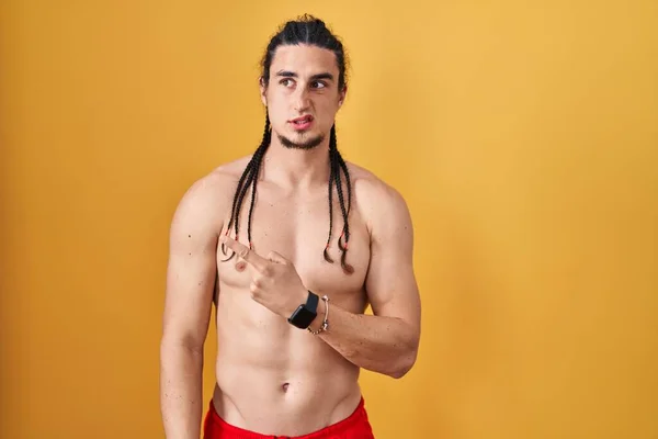 Hispanic man with long hair standing shirtless over yellow background pointing aside worried and nervous with forefinger, concerned and surprised expression