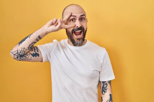 Young hispanic man with tattoos standing over yellow background doing peace symbol with fingers over face, smiling cheerful showing victory