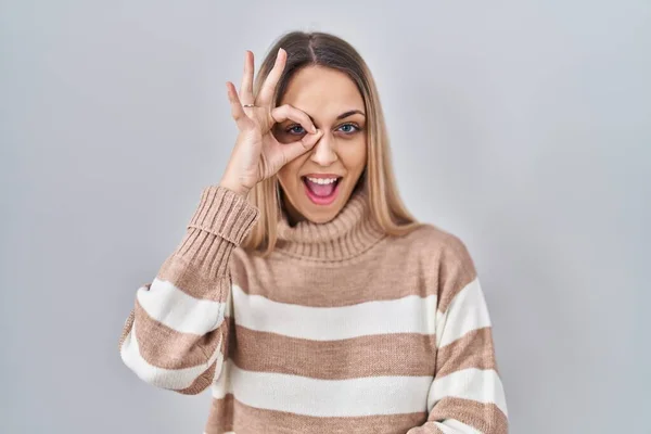Young blonde woman wearing turtleneck sweater over isolated background doing ok gesture with hand smiling, eye looking through fingers with happy face.