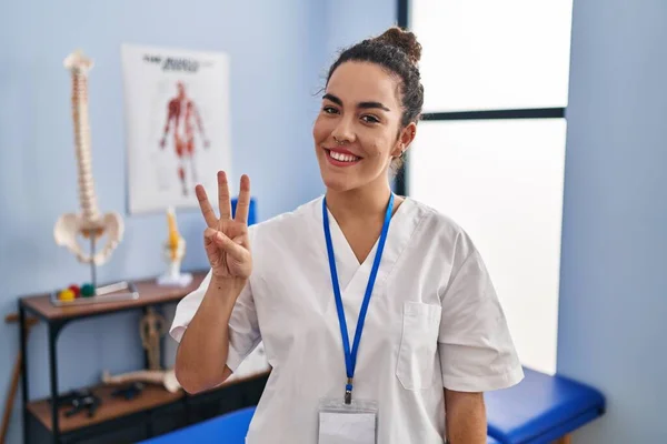 リハビリクリニックで働く若いヒスパニック系の女性が自信と幸せな笑顔で指の数3を見せ 指差し込み — ストック写真