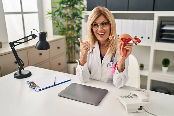 Middle age blonde gynecologist woman holding anatomical model of female genital organ pointing thumb up to the side smiling happy with open mouth